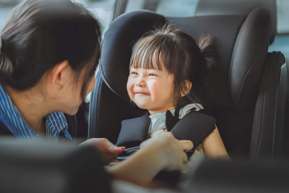 A,Loving,Asian,Mother,Carefully,Buckles,Her,Smiling,Toddler,Daughter child in car seat. road trip safety tips for kids